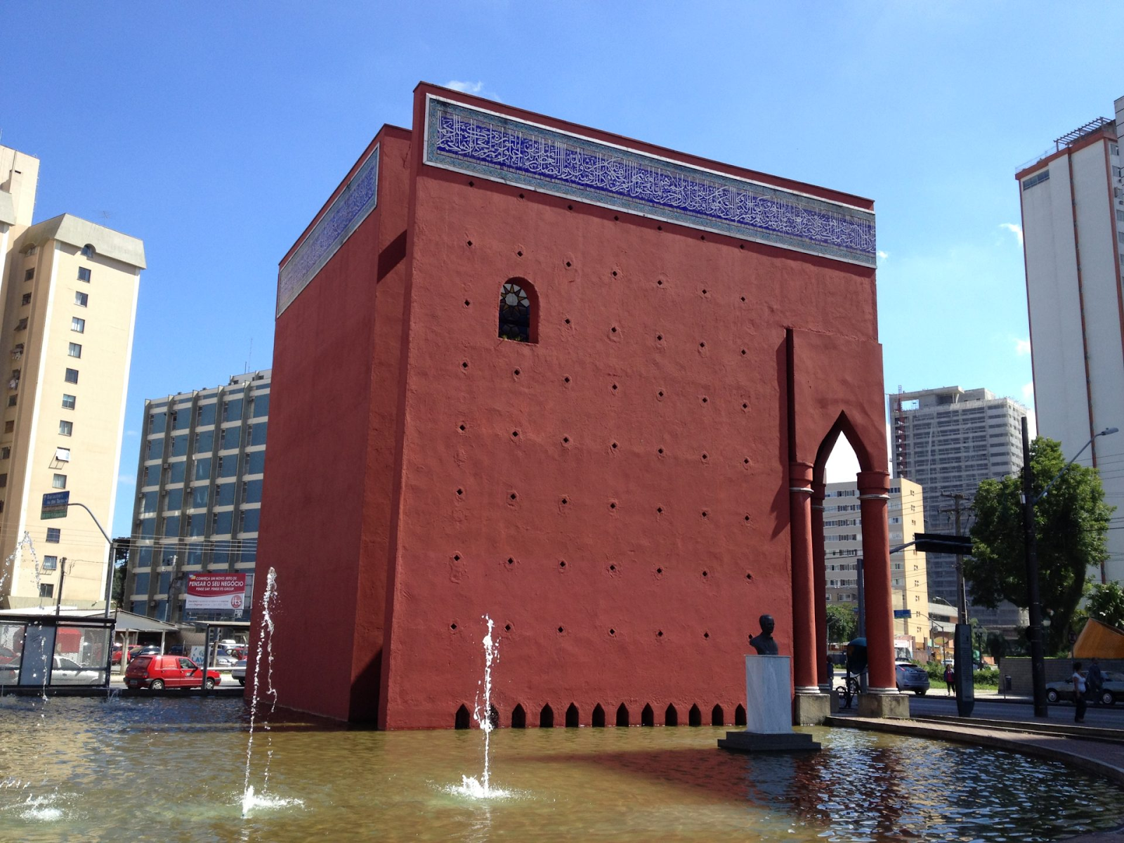 Edifício vermelho com fachada lisa, janelas pequenas e detalhes arquitetônicos de influência islâmica, localizado no centro de uma praça urbana cercada por prédios altos. O edifício está parcialmente rodeado por um espelho d’água com fontes verticais e uma escultura de busto em pedestal próximo à entrada. O céu está claro e azul, indicando um dia ensolarado.