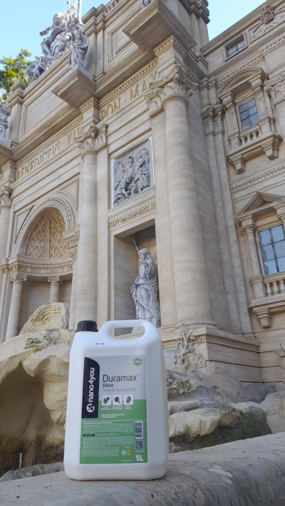 Prédio histórico de estilo clássico com colunas romanas, esculturas e detalhes arquitetônicos esculpidos em pedra clara, remetendo à Fontana di Trevi, em Roma. Em primeiro plano, aparece parcialmente um galão branco de produto de limpeza com tampa preta, contrastando com a monumentalidade da construção ao fundo. A imagem sugere o uso do produto em contextos de preservação ou limpeza de superfícies históricas ou artísticas.